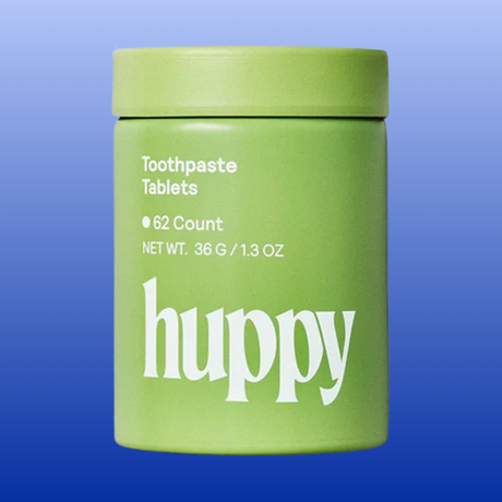A light green cylindrical container of Huppy Toothpaste Tablets on a blue gradient background. The container features white text with the brand name 'huppy' in a stylized lowercase font.